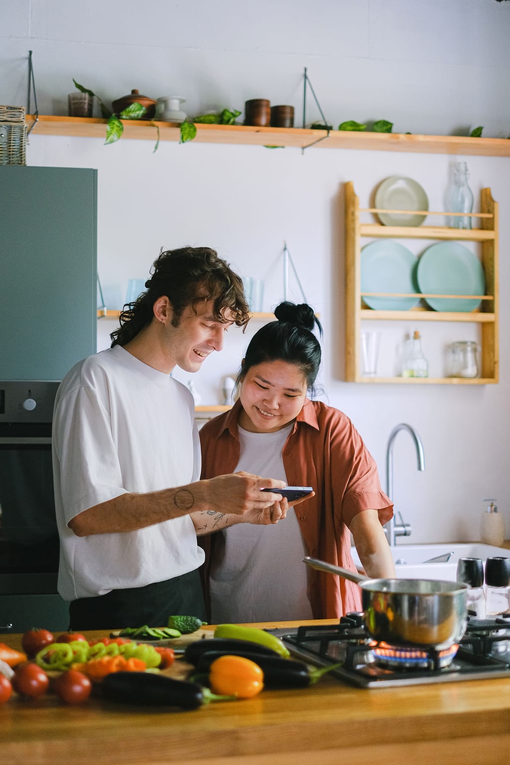 Two people in a kitchen look at a phone together, vegetables chopped on the bench