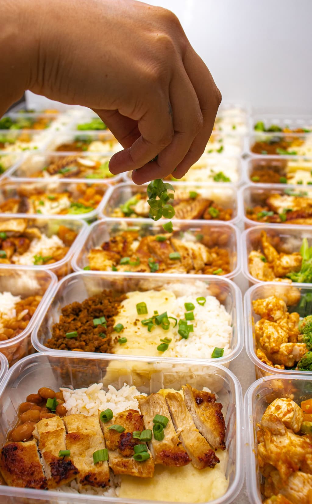 A hand sprinkles spring onion over rows of meal-prep containers of chicken, rice and mince